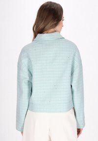 Sacou de tweed bleu deschis cu un model texturat, în rețea. Prezintă un guler și o croială lejeră, cu un tiv scurt și mâneci lungi.