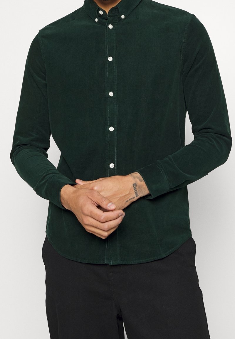 Homme portant une chemise en velours côtelé vert foncé à boutons blancs, pantalon noir, les mains jointes montrant un tatouage au poignet.