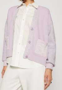 Lila stickad cardigan med ribbad textur, med knappstängning, två fickor och kontrasterande ärmdesign. Bärs över en vit skjorta.