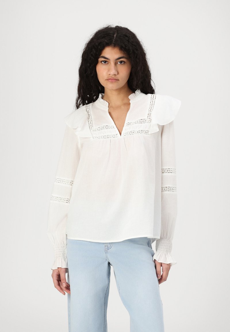 Blusa bianca con collo arricciato, dettagli in pizzo sul petto e sulle maniche, e polsini smock. Abbinata a jeans blu chiaro.