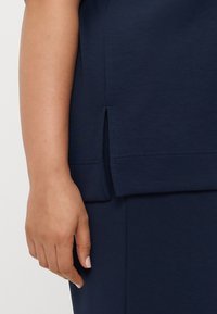 Ναυτικό μπλε μπλούζα με πλευρική σχισμή στο τελείωμα, φτιαγμένη από μαλακό ύφασμα, με ορατές ραφές. Ένα χέρι ακουμπά στην επιφάνεια.