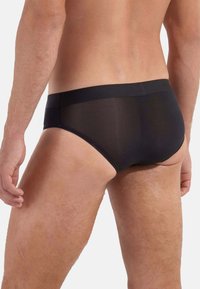 Schwarze Herrenunterwäsche mit einem glatten elastischen Bund und transparentem Stoff. Sie verfügt über ein enganliegendes Design und minimale Abdeckung am Rücken.