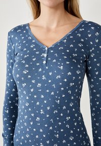 Top blu lungo a maniche, realizzato in tessuto testurizzato con motivo floreale. Presenta un colletto a V e una chiusura con tre bottoni.