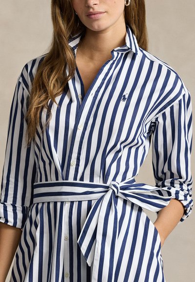 Nő, aki tengerészkék és fehér, függőleges csíkos ingruhát visel, megkötött derékövvel és feltűrt ujjakkal, áll, egyik kezét a zsebében tartva.