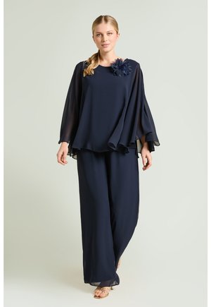 Frau trägt eine marineblaue, fließende Bluse mit transparenten langen Ärmeln und passenden weit geschnittenen Hosen, dazu eine marineblaue Blumenbrosche.