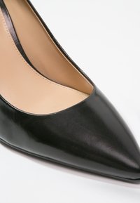 Chaussure à talons hauts en cuir noir avec un bout pointu et une finition lisse. L'intérieur est de couleur nude avec des détails de couture minimalistes.