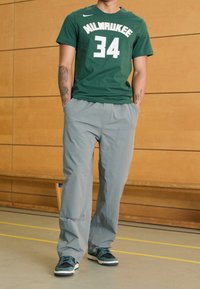 Zelené bavlněné tričko s nápisem "MILWAUKEE 34" v bílé barvě, doplněné o světle šedé volné kalhoty a černé tenisky s tóny tyrkysové.