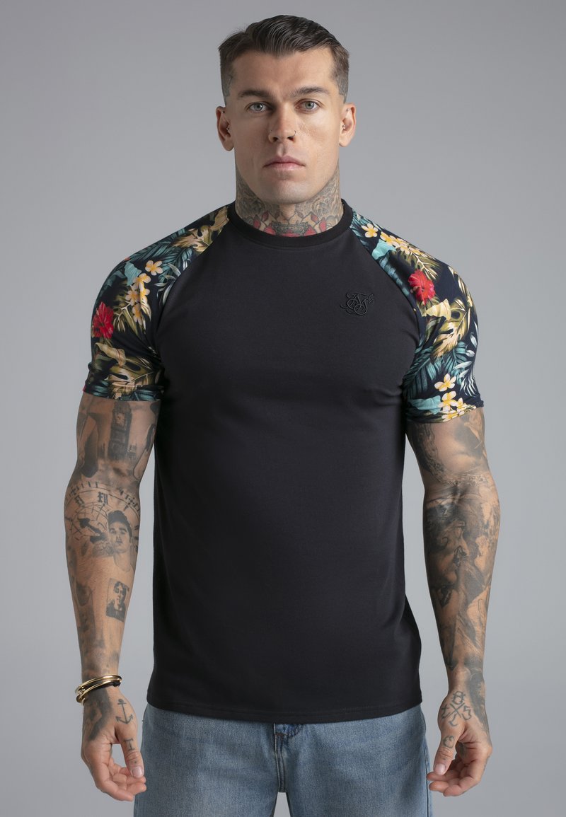 Uomo con capelli corti e tatuaggi visibili indossa una maglietta nera con motivo floreale sulle maniche e jeans azzurri, in piedi davanti a uno sfondo semplice.