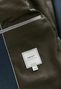 Schlank geschnittene Tailor-Jacke in dunklem Oliv, mit einem subtilen geometrischen Muster, grauen Akzenten und einem weißen Etikett mit Markendetails.
