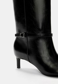 Stivale in pelle nera alto fino al ginocchio, con punta affusolata, tacco sottile e una cintura decorativa attorno alla caviglia. Texture liscia e finitura lucida.