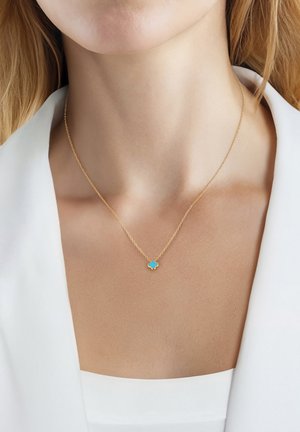 Delicado collar de cadena de oro con pequeño colgante de trébol de cuatro hojas azul, llevado en el cuello de una mujer sobre ropa blanca.