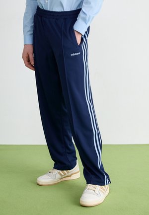 Person iført marineblå Adidas træningsbukser med hvide striber og logo, kombineret med hvide og beige sneakers, stående på grøn gulv.