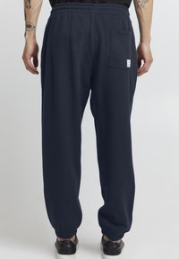 Marineblå joggebukser med elastisk midjebånd, mansjetter på anklene og en baklomme med en liten merkeetikett. Laget av bomull.