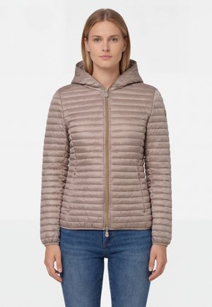 Donna che indossa una giacca trapuntata taupe con cappuccio e cerniera frontale, e jeans blu, in piedi contro uno sfondo bianco.