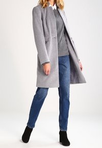 Abrigo gris de mezcla de lana con corte recto, dos bolsillos frontales y solapa. Combinado con un suéter gris, vaqueros azules y botas negras hasta el tobillo.