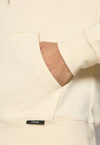 Kremo barve kapuca s kenguru žepom, ki ima črno oznako "CITY EDITION". Mehka tekstura in rebrasti manšeti izboljšujeta njegov dizajn.