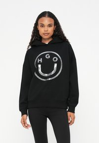 Czarna bluza z kapturem z błyszczącym srebrnym okrągłym logo zawierającym napis "HGO" oraz uśmiechniętą buźkę. Umieszczona na gładkim tle, o miękkiej teksturze.