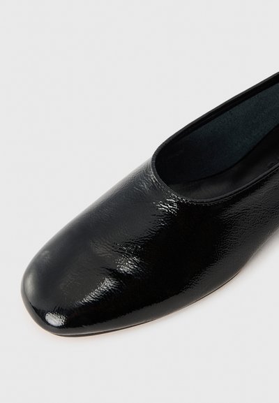 Chaussure slip-on en cuir verni noir avec un bout arrondi, texture lisse, design minimaliste et silhouette basse.