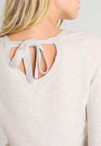 Lys beige striktrøje med hjerteformet udskæring bagpå, bundet med matchende stof. Blød tekstur, afslappet design.