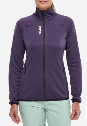 Femme portant une veste softshell violette zippée avec des poches latérales zippées et un pantalon vert clair, vue du cou jusqu'aux hanches.