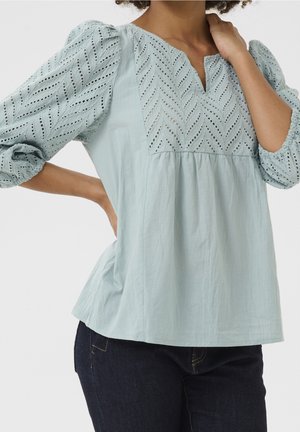 Donna che indossa una camicetta azzurra chiaro a occhielli con maniche a sbuffo tre quarti e jeans blu scuro, posando con una mano sul fianco e l'altra vicino al collo.