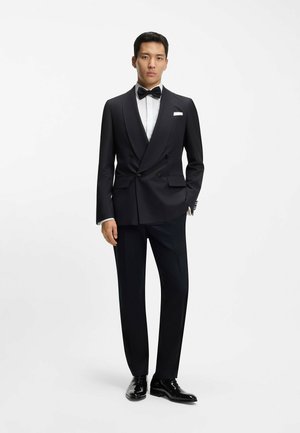 Un homme se tient vêtu d'un smoking noir à revers châle, d'une chemise blanche, d'un nœud papillon noir, de chaussures en cuir verni noir et d'un mouchoir de poche blanc.