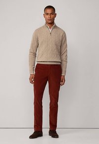 Beige kabelgebreide trui met een kwartritssluiting bij de kraag, gecombineerd met roestbruine ribfluwelen broek. Donkerbruine schoenen, staand tegen een neutrale achtergrond.