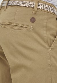 Pantalonii khaki au un design curat, cu o textură netedă, o buzunar în spate și o etichetă cu logo maro. Talie este decorată cu accente albe și maro.