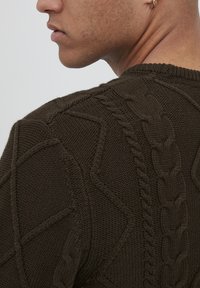 Pulover tricotat verde închis, cu modele de tip cable knit și geometrice, cu guler rotund ribanat și detalii texturate pe țesătură.