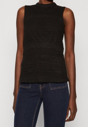 Haut sans manches en dentelle noire à col montant porté avec un jean bleu foncé, montrant la texture détaillée du tissu et des poches cousues.