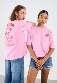 Zwei Personen tragen leuchtend pinkfarbene übergroße T-Shirts. Auf der Rückseite eines Shirts befindet sich eine mehrfarbige Schmetterlings- und Blumen Grafik. Eine Person trägt blaue Jeansshorts.