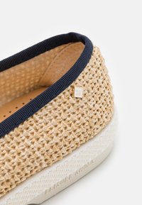 Vlechbanden met een tan textiel bovenkant en marineblauwe accenten, met een klein logo detail. Getextureerde witte zool met een subtiel patroon aan de rand.