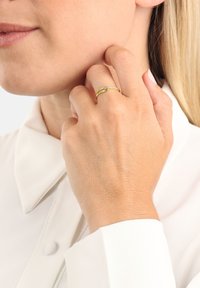 Närbild av en person som bär en ring av guld och diamanter på fingret, som rör vid sin kind, klädd i en vit skjorta med krage.