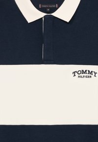 Sötétkék és fehér poloing, texturált anyagból készült. Klasszikus gallérral, gombos záródással és hímezett "Tommy Hilfiger" logóval rendelkezik.