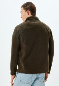 Chaqueta de forro polar verde oscuro con un alto cuello y mangas raglán, que presenta una textura suave y un diseño ajustado. Costuras visibles a lo largo de las uniones.