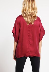 Röd satinblus med vida ärmar, en flödande form och en mittbaksöm. Den släta texturen förstärker dess lyxiga utseende.