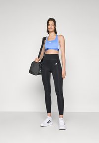 Blå sportsbh med hvide striber, sorte højtaljede leggings og hvide sportssko. Model bærer en sort træningstaske.