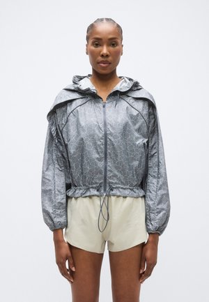 Ľahká sivá vetrovka s textúrovaným vzorom, nastaviteľnou kapucňou a zúženým pásom. Kombinovaná s krémovými šortkami s sieťovými prvkami.