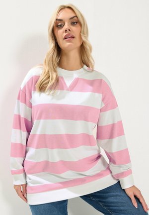 Femme aux cheveux longs et blonds portant un sweat-shirt ample à rayures horizontales roses et blanches ainsi qu'un jean bleu, sur fond blanc.