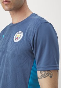 Mančestras City krekls zilos toņos ar teksturētu rakstu, ar logotipu uz krūšu daļas un tirkīza sānu akcentiem. Īsas piedurknes, sportisks piegriezums.