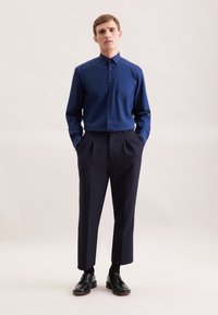 Lange mouwen, aansluitend donkerblauw overhemd met knopen en een borstzak, gecombineerd met hooggesneden, geplooide donkere broek en zwarte nette schoenen.