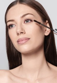 Brązowa szczoteczka do tuszu do rzęs trzymana blisko oka, z gładką cerą i prostymi włosami. Tło jest jasnoszare, co podkreśla główny temat zdjęcia.