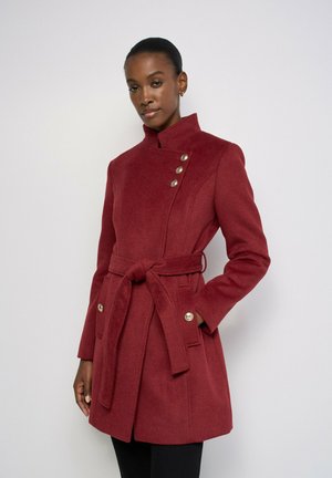 Femme portant un manteau rouge longueur genou avec une ceinture à la taille, un col montant et trois boutons sur la poitrine et les poches, debout devant un fond blanc.