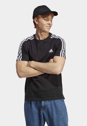 Zwarte t-shirt met een witte drie-strepen ontwerp op de schouders en een klein Adidas-logo. Gekozen in combinatie met blauwe spijkerbroek en een zwarte pet.