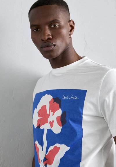Balta T-krekls ar lielu ziedu grafiku sarkanā, zilā un melnā krāsā. Signatūras "Paul Smith" uzraksts augšējā stūrī. Gluda tekstūra.