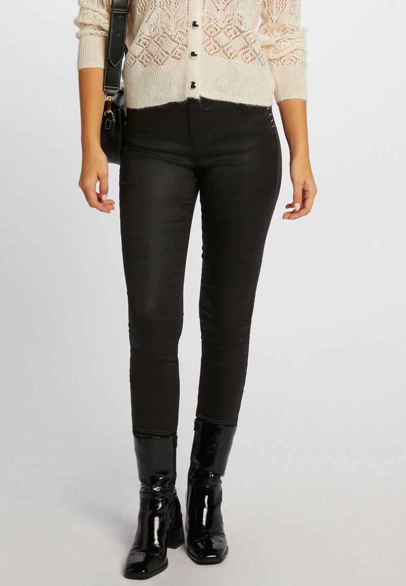Femme portant un pantalon skinny noir, des bottines en cuir verni noir, un cardigan en maille crème et portant un sac à bandoulière noir.