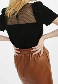 Svart topp med ett nät bakpanel, puffärmar och bruna byxor med elastisk midja, som har en slät yta och lös passform.