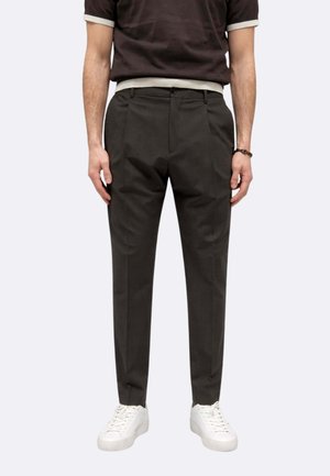 Uomo con pantaloni neri su misura, sneakers bianche e camicia nera a maniche corte con bordi bianchi, in piedi davanti a uno sfondo chiaro.