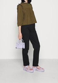 Olivegrüne Bluse mit Struktur und Rüschensleeves, kombiniert mit schwarzen Hosen, einer lavendelfarbenen Mini-Handtasche und klobigen lavendelfarbenen Sandalen mit Riemen.