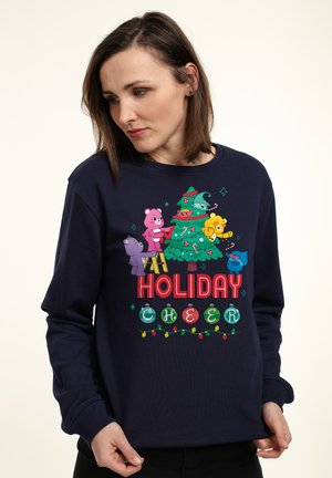 Marineblåt sweater med et farverigt grafisk motiv af tegneserie-dyr, der dekorerer et juletræ med pynt og lyskæder; teksten siger "HOLIDAY CHEER."